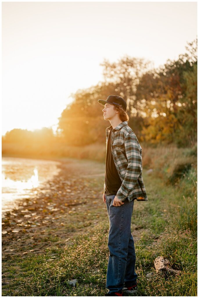 the golden sun shines out from behind trees as boy looks out over water during a senior portrait session for your teen