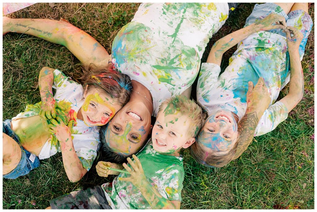 mom lays on grass with kids and all are covered in paint by Minnesota wedding photographer
