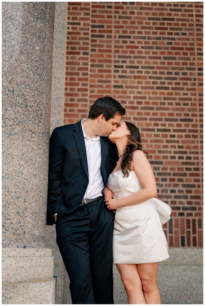 couple kisses as man leans against column at Downtown St. Cloud engagement session