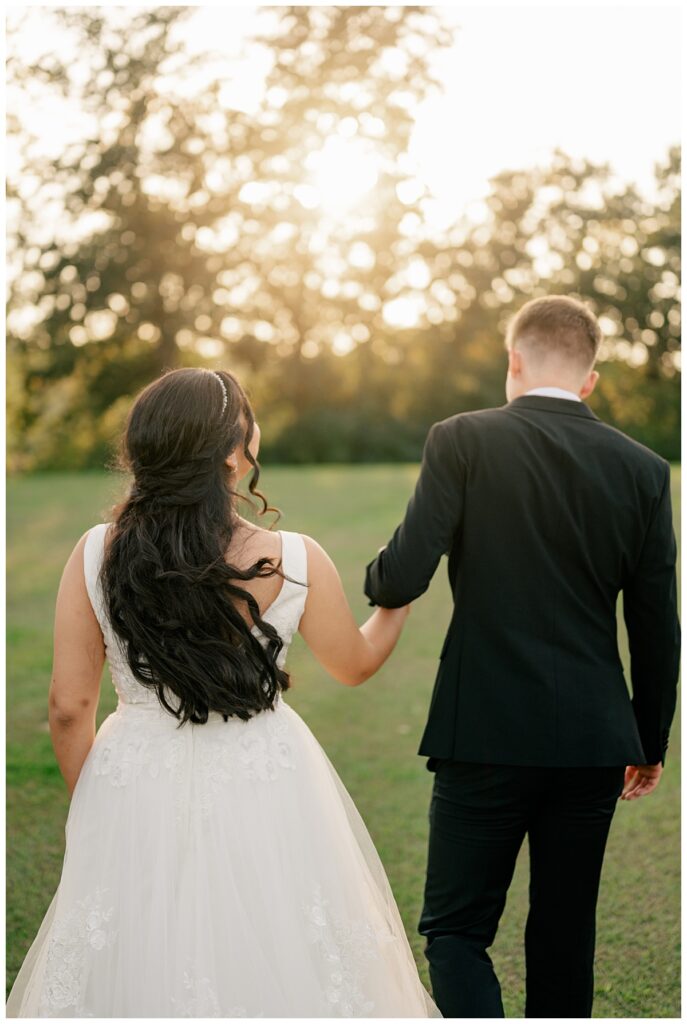 groom holds bride's hand as they walk toward the sunset during golden hour by Rule Creative Co