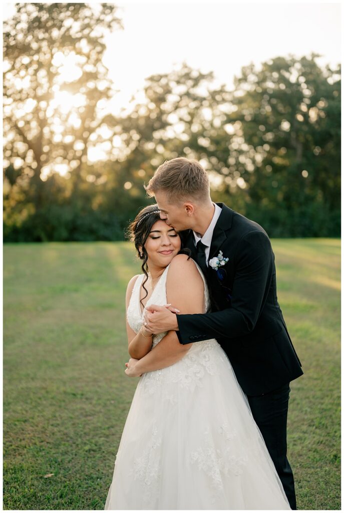 man wraps arms around woman from behind and kisses her forehead at late summer wedding in Paynesville