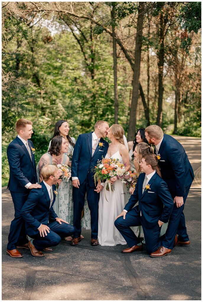 wedding party gathers around as bride and groom kiss by Rule Creative Co