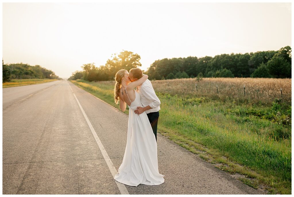 man and woman kiss on road as sun sets behind them after Northern Oaks summer wedding