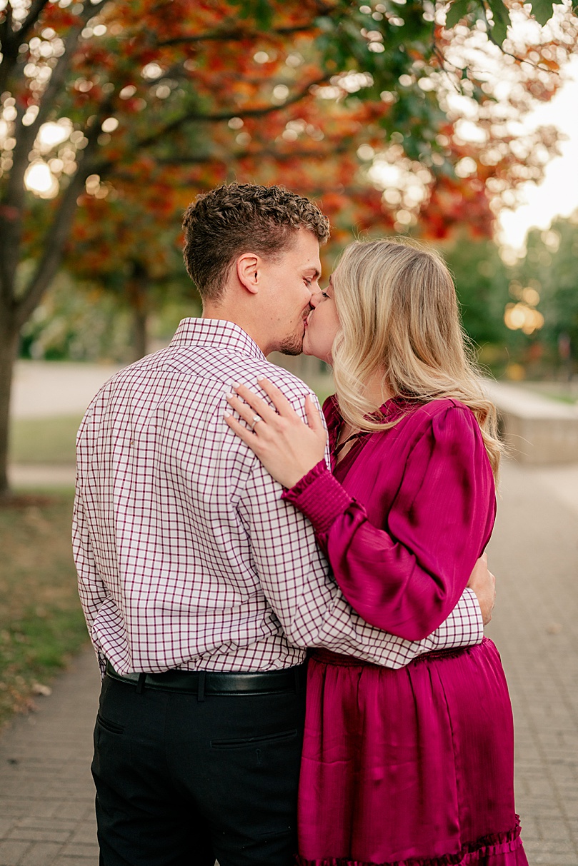 man kisses fiance under trees with fall colors by Minnesota wedding photographer