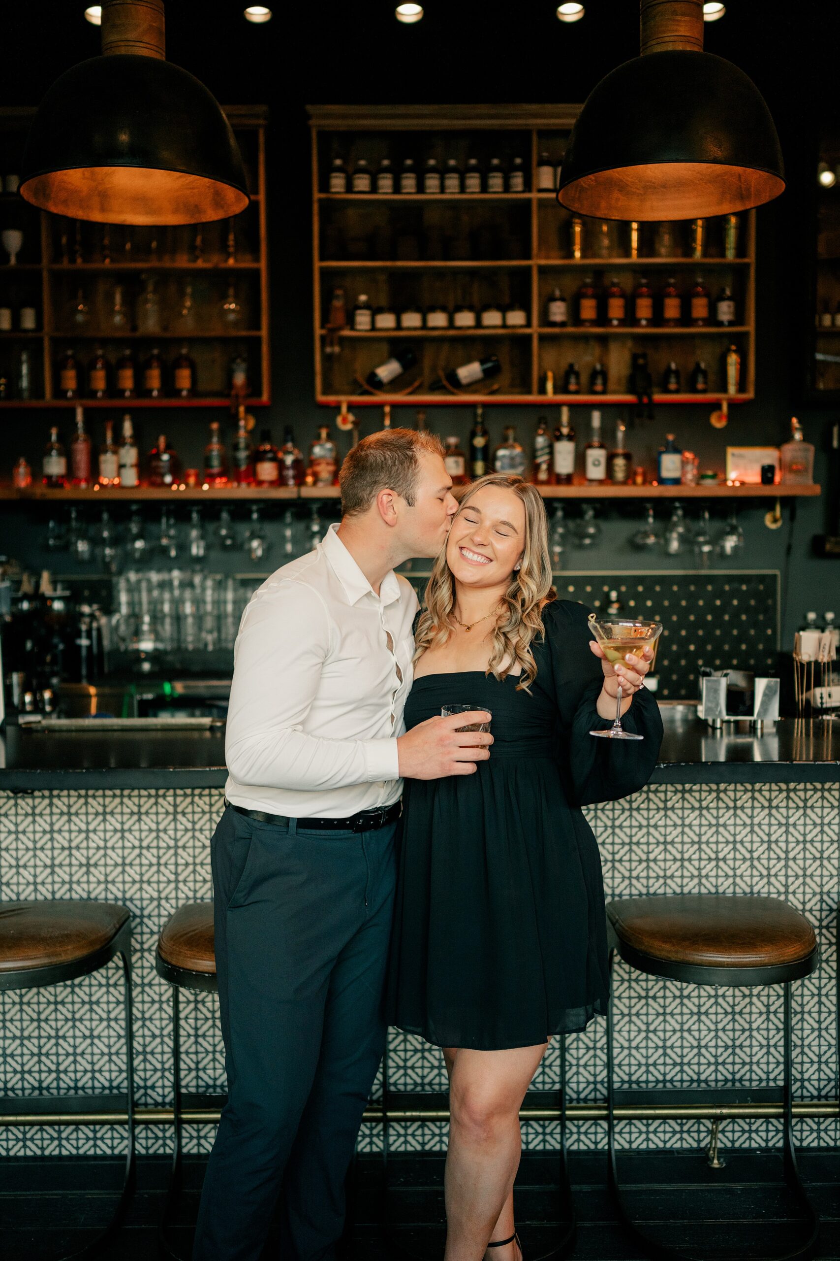 Man kisses fiance at bar holding cocktails by Minnesota Wedding Photographer