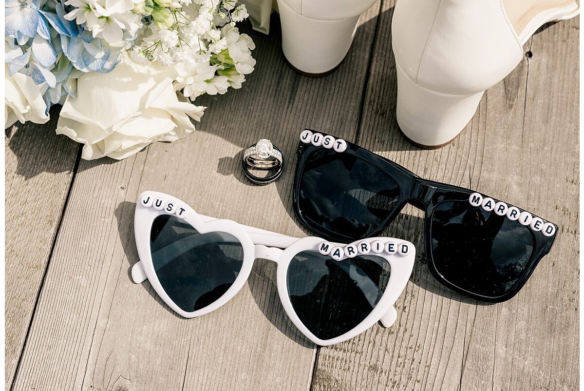 An aerial photograph of a wedding bouquet, white chunky high heels, wedding rings, and 2 pairs of sunglasses that read "Just Married" across the frames.