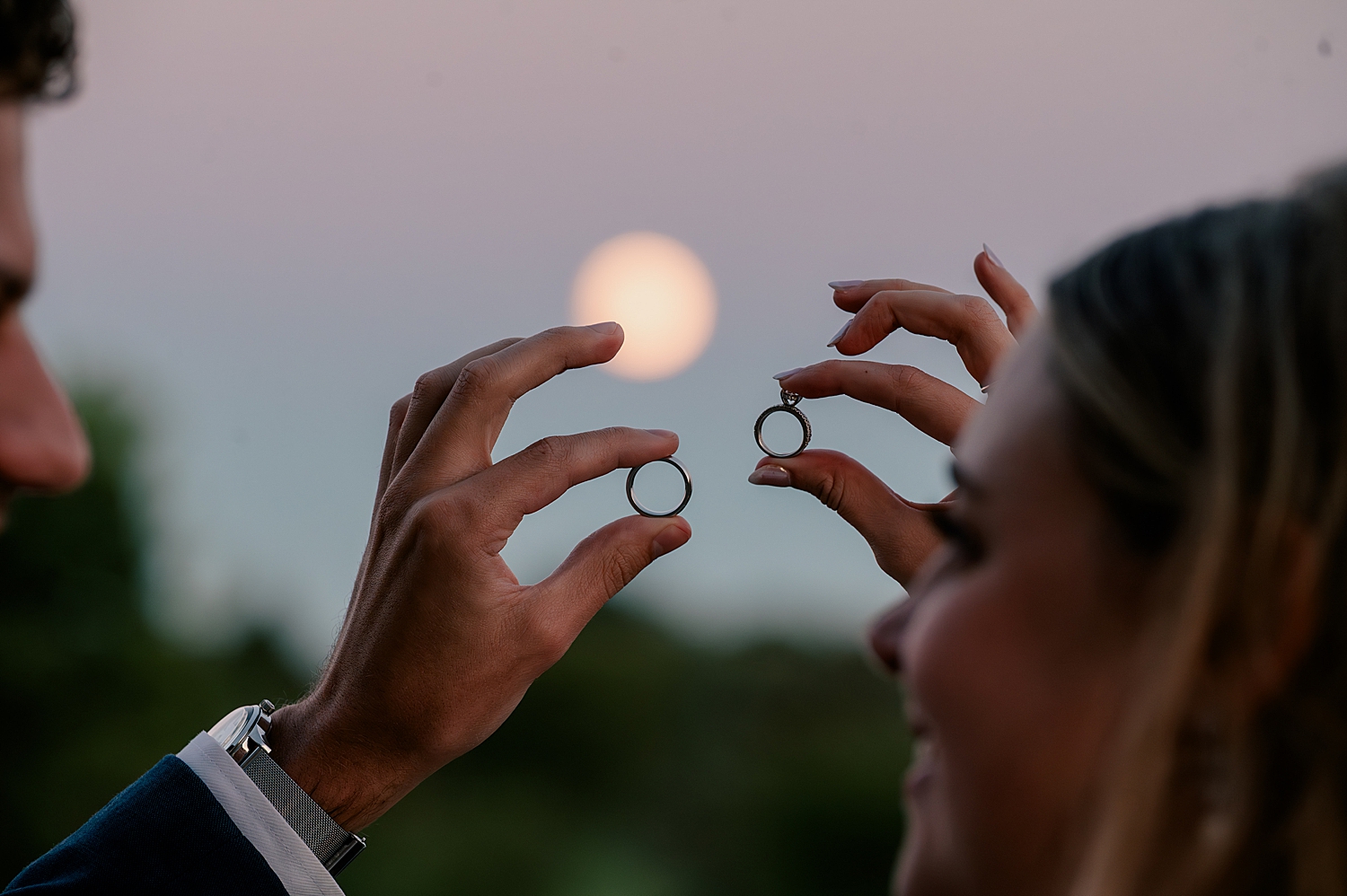 man and wife hold up their rings in front of an emerging moon at sunset by Rule Creative Co