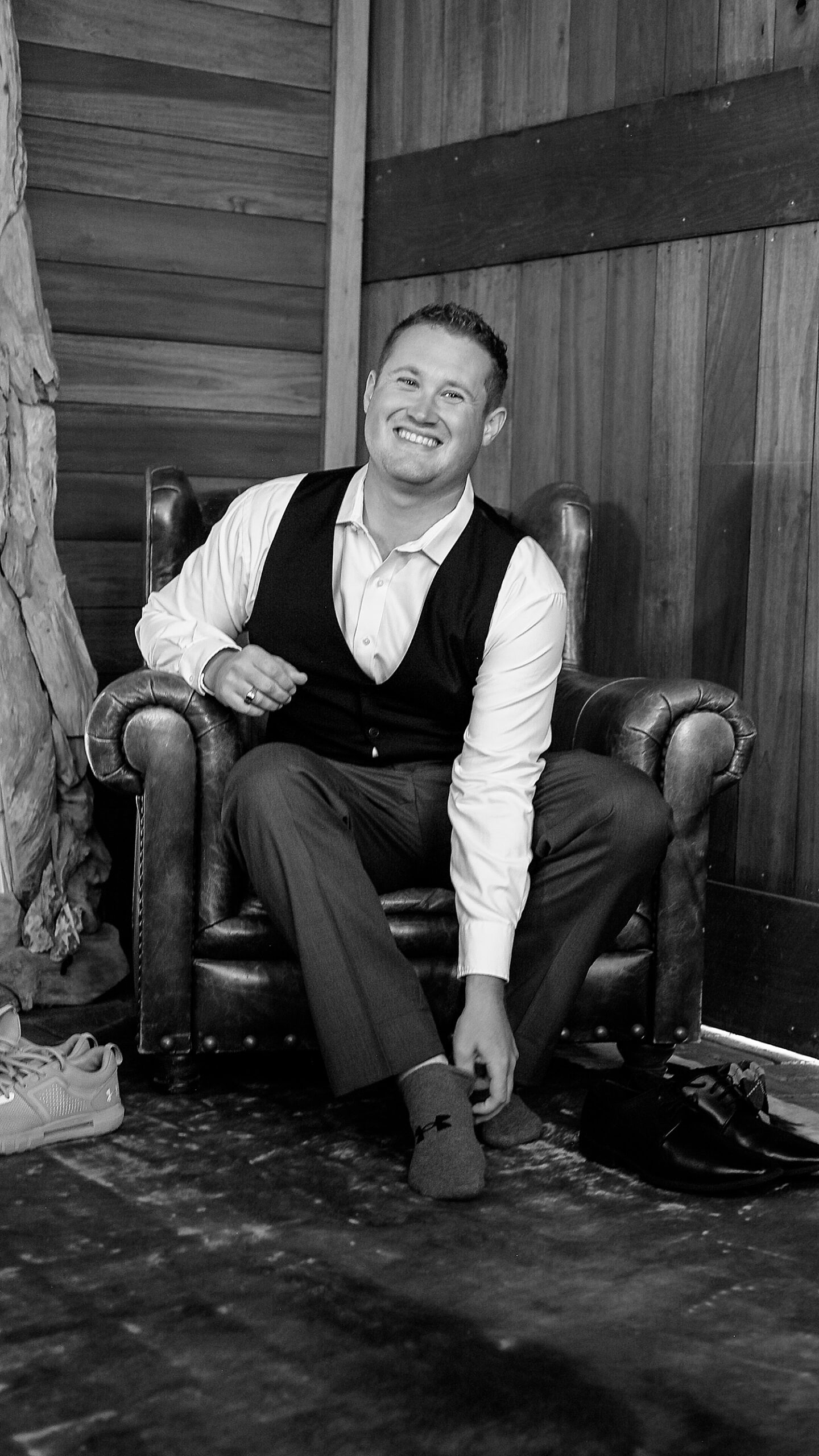 groom grins in leather chair, putting on his shoes before big day at Redeemed Farm venue