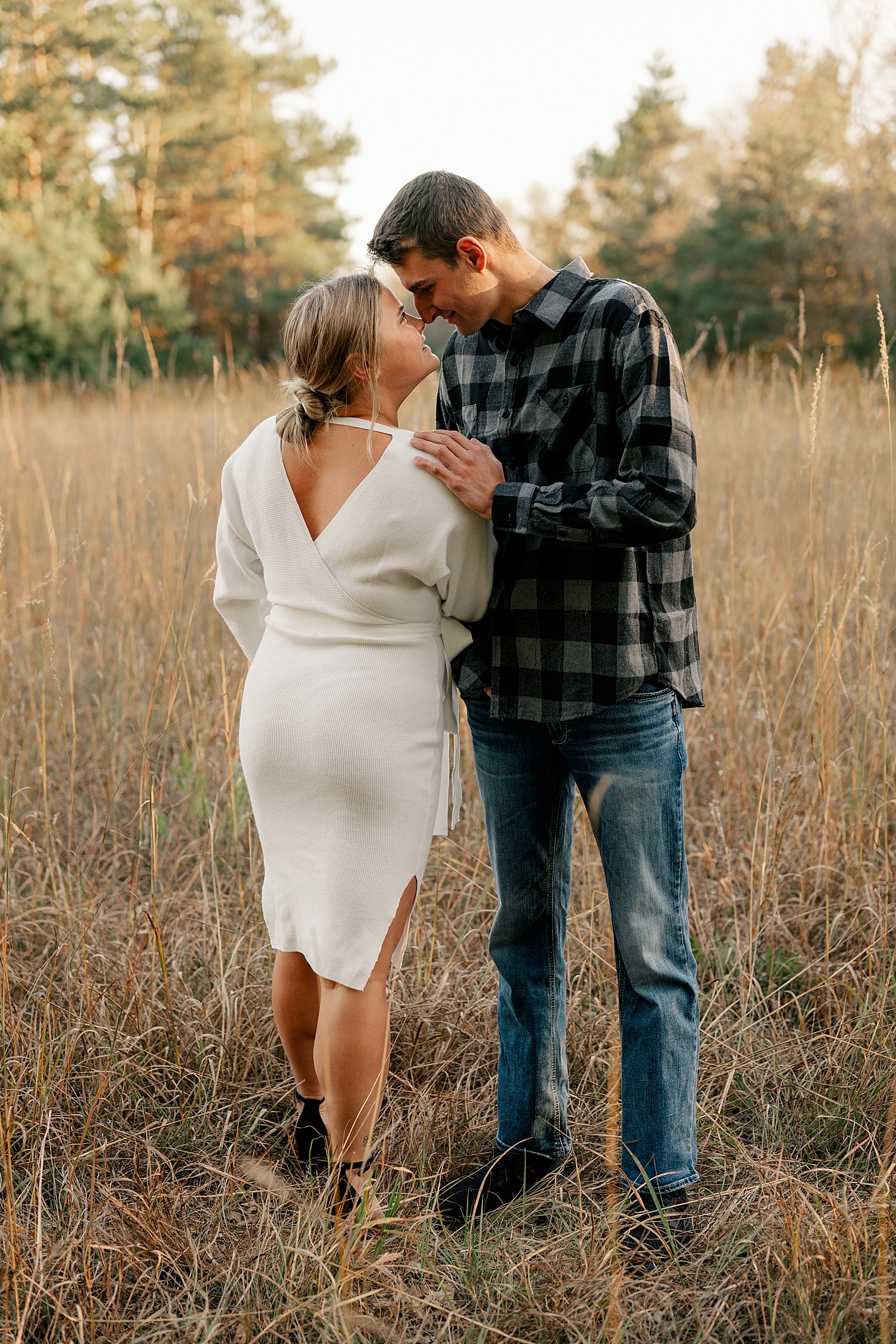 man in plaid looks down at his wife-to-be in field for sunset engagement session