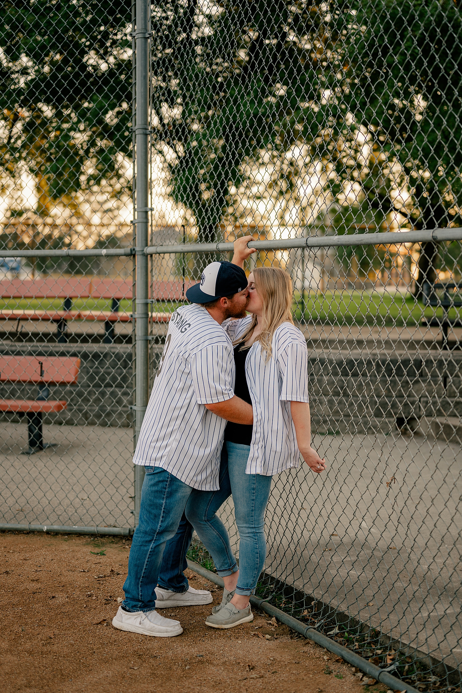 man leans into his fiance against the metal fence for engagement session at baseball field