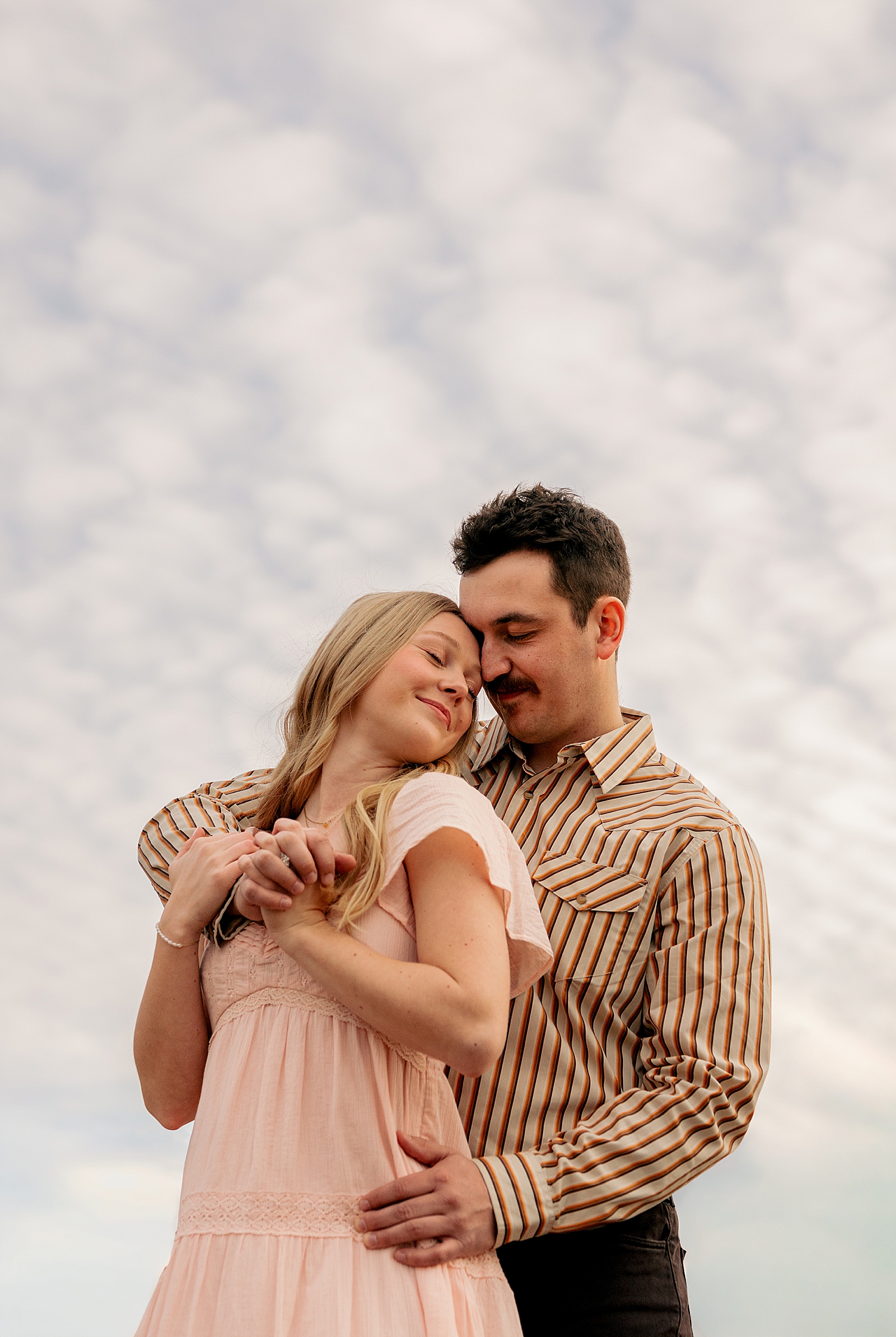 engaged couple stand in field with clouds behind them for playful engagement session