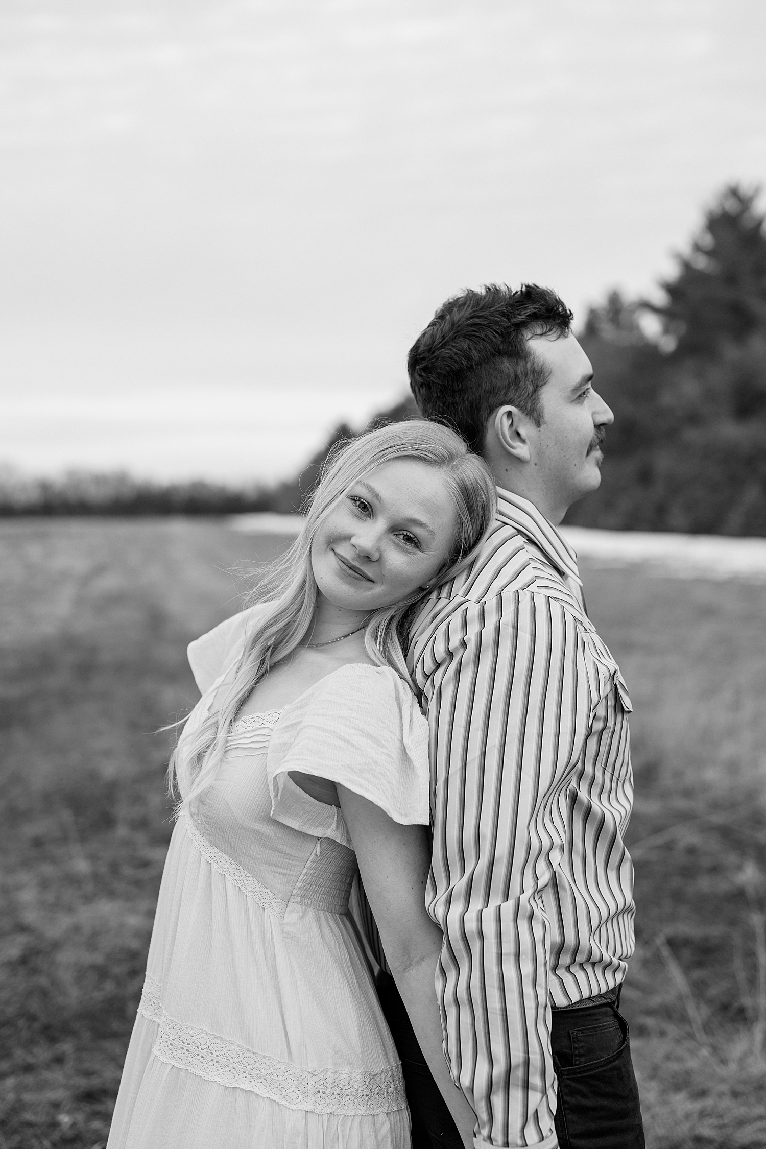 man and woman stand back to back in field during playful engagement session