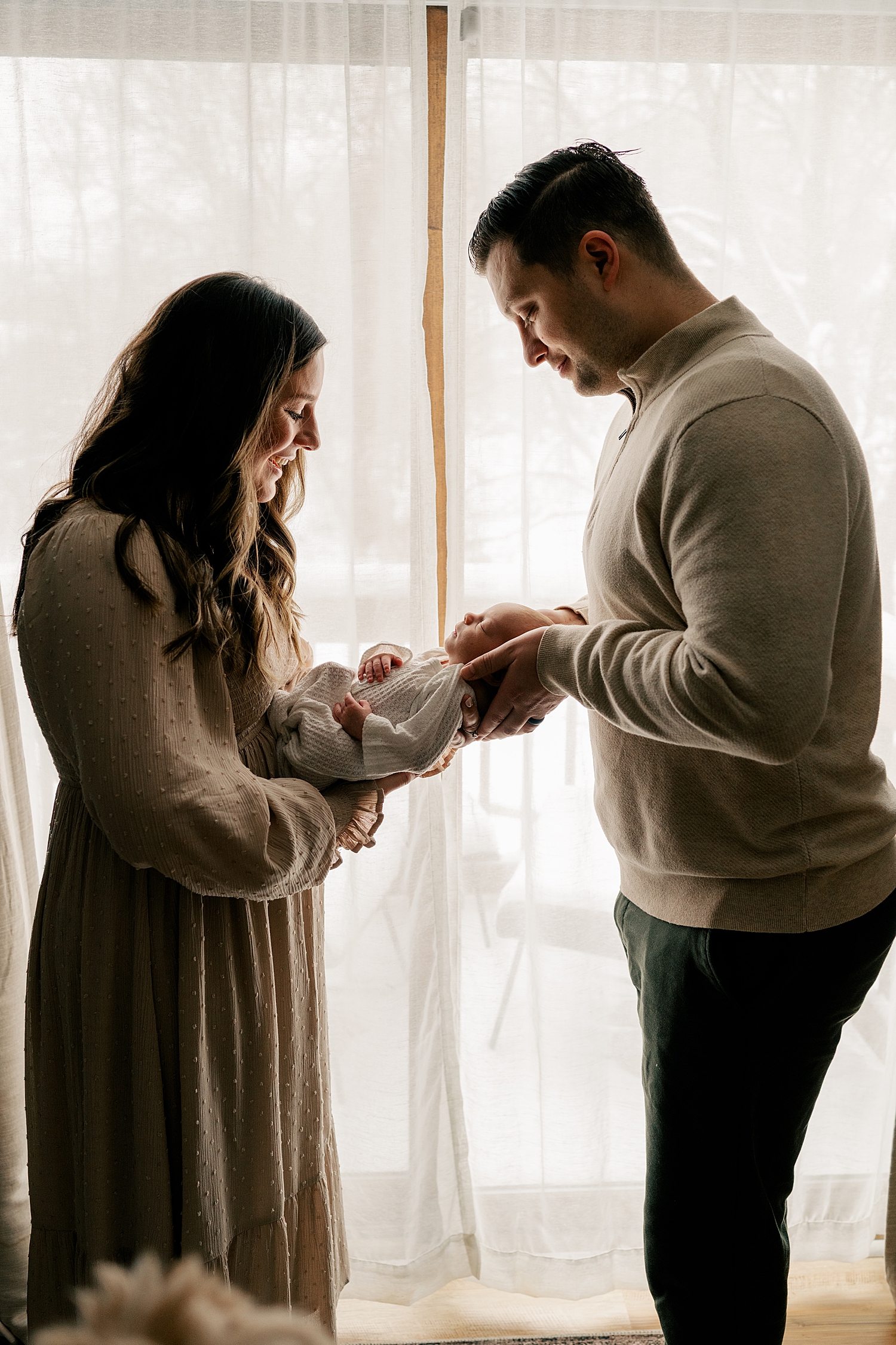 mom and dad stand in front of window holding out their new addition by Rule Creative Co