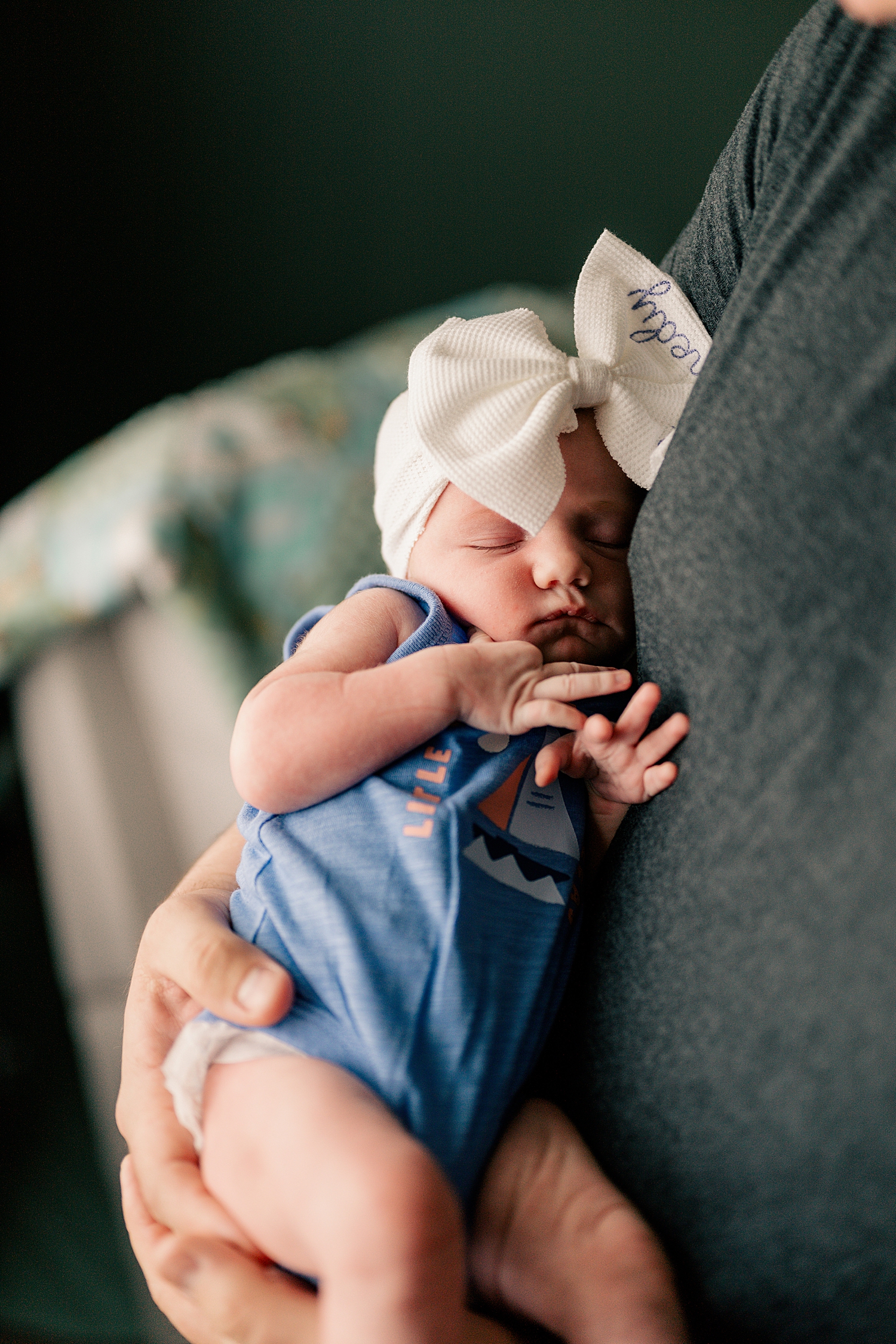 little baby in blue onesie and bow is held by her father for in-home session