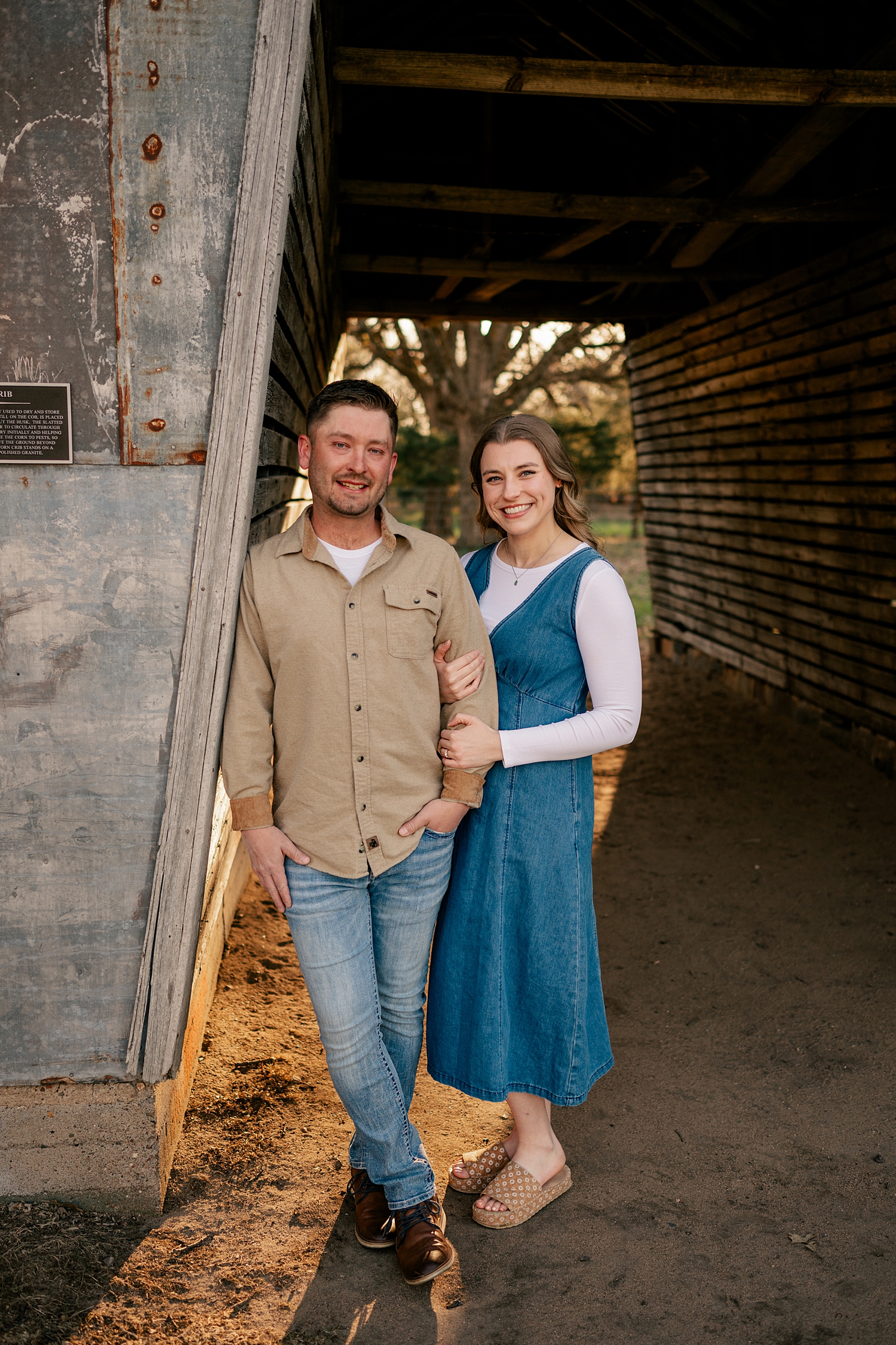 woman in denim dress and man in tan shirt lean on barn for summer engagement photos