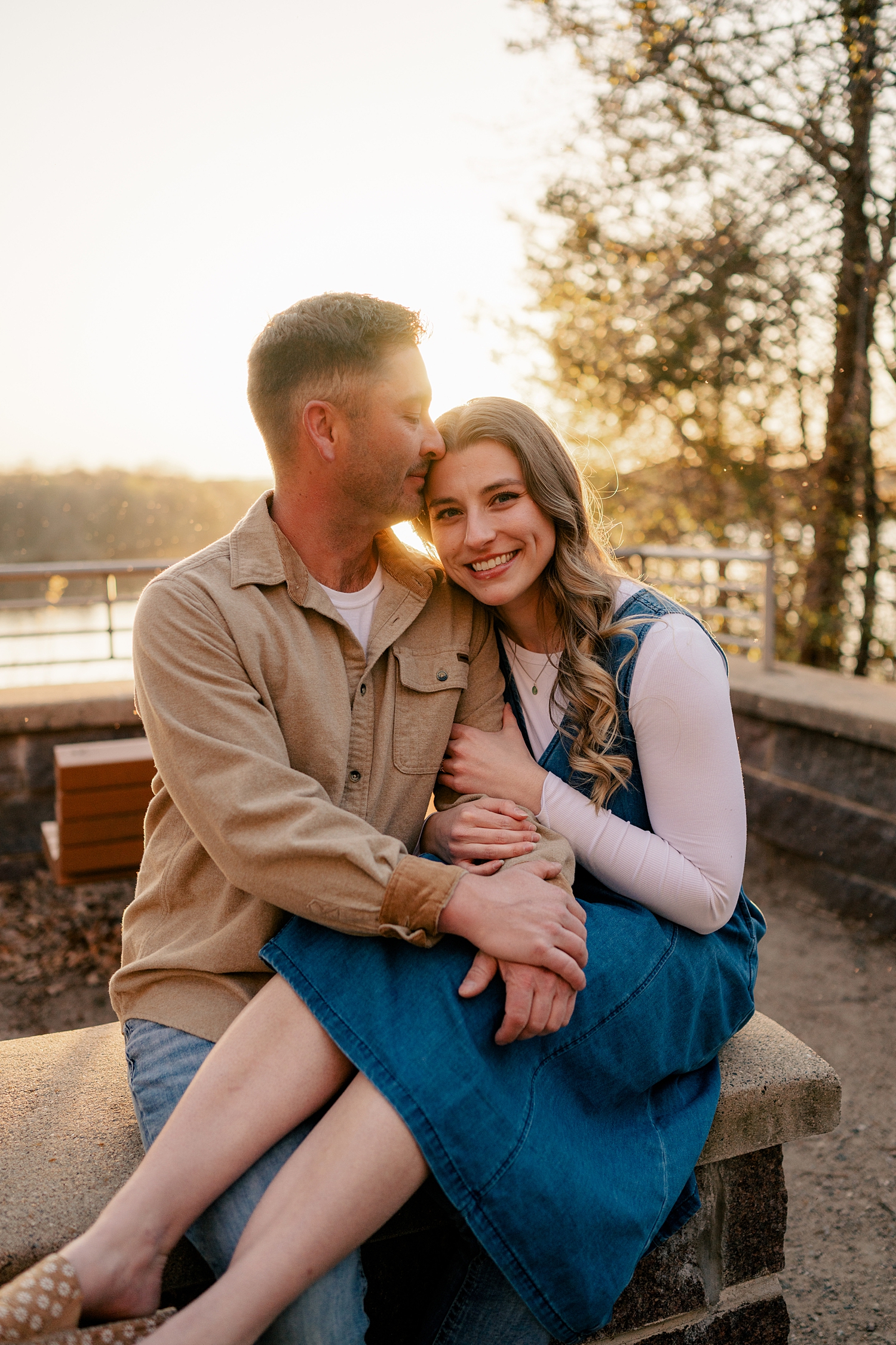 groom to be leans in to kiss his fiancé's temple as they sit on stone bench by Rule Creative Co